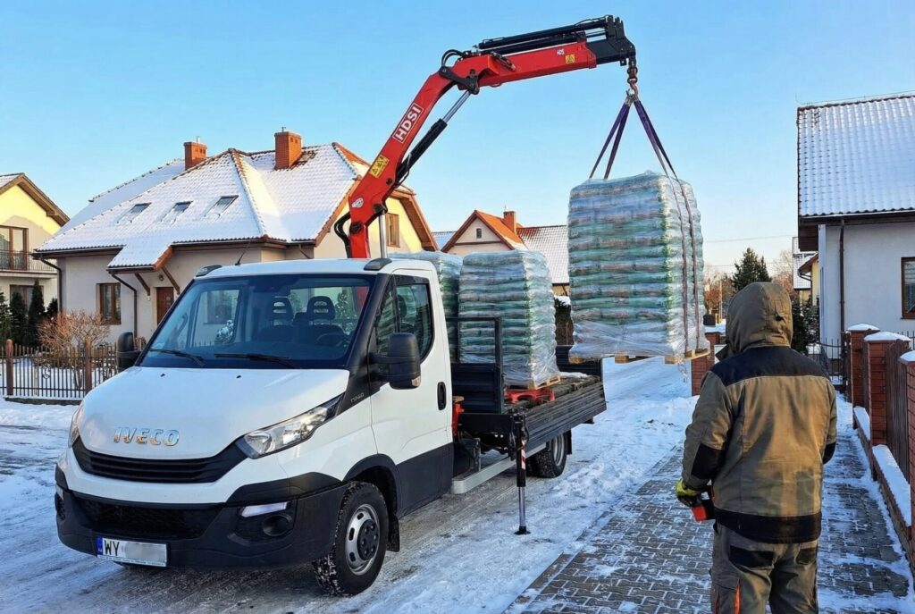 Halinów - Dostawa pelletu drzewnego za pomocą wózka widłowego w zimowej scenerii. Mężczyzna obserwuje podnoszenie palet z pelletem, co ilustruje proces transportu tego ekologicznego materiału opałowego.